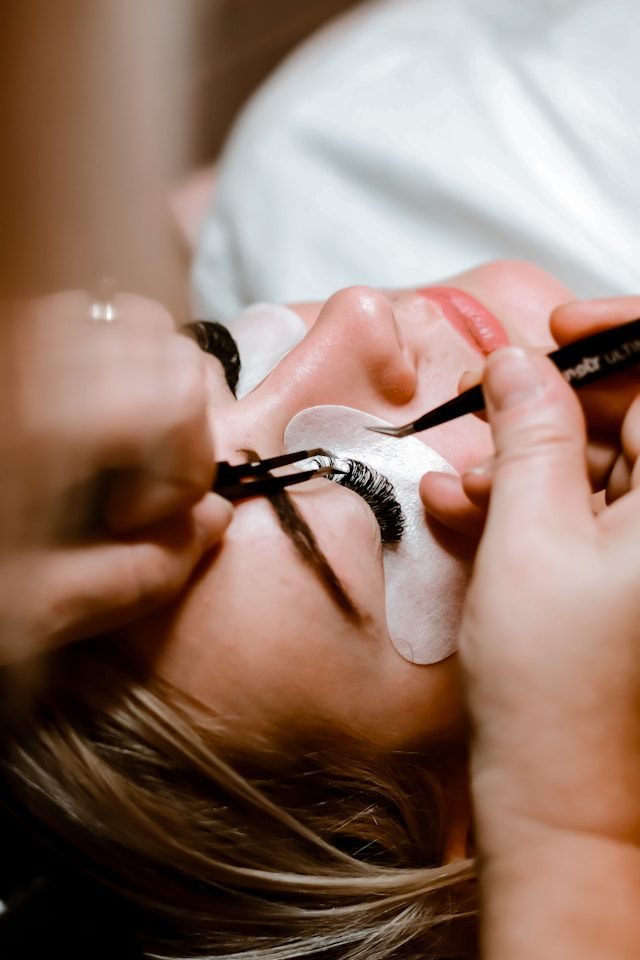 A woman laying on a bed, she has felt padding under her eye getting her eyelashes placed