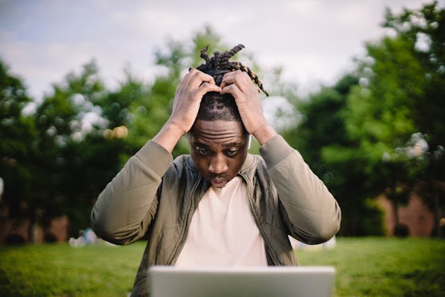 A man grabbing his head in dismay looking at his laptop in a park