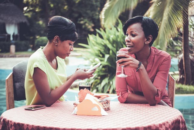 2 black women, one scrolling through her phone and the other drinking a glass of red one deep in thought