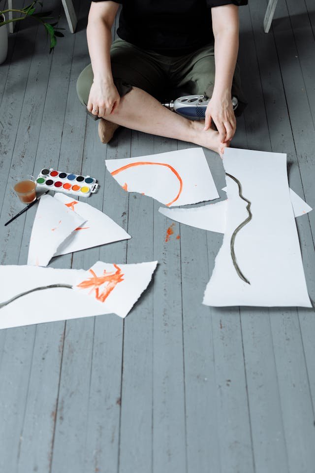 A man sitting on the floor with a ripped up painting and a set of paints next to it. 