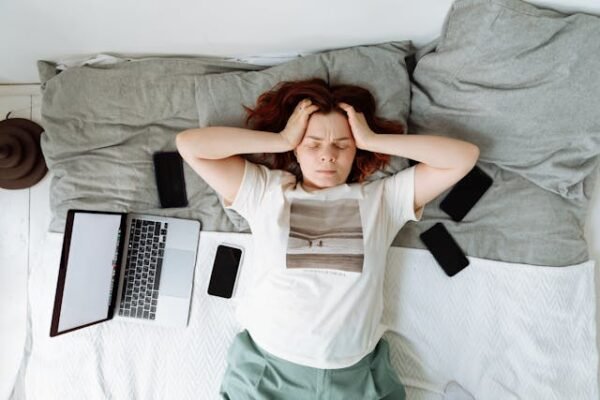 A woman laying on her back on a bed with her hands on each side of her head looking in dispair with an open laptop and several mobile phones by her side