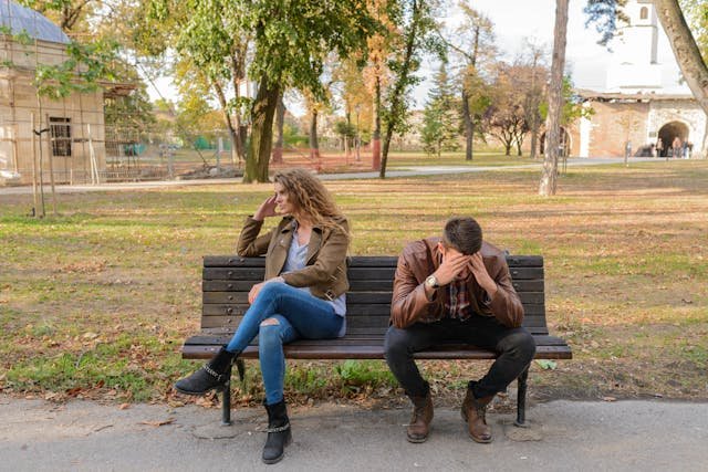 Image of a woman and a man sitting on a brown bench looking upset and disconnected

