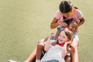 A row of 3 kids on a grass, tickling and playing with each other smiling and happy.